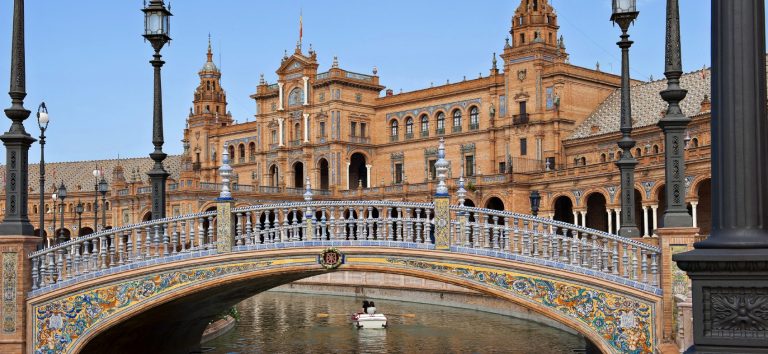 Puente sobre el canal frente a un majestuoso edificio de estilo regionalista en Sevilla.