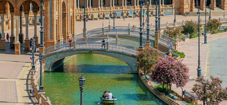 Puente sobre el canal frente a un majestuoso edificio de estilo regionalista en Sevilla.